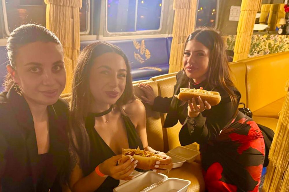 Three women sitting on a yellow couch holding hot dogs, smiling at the camera.