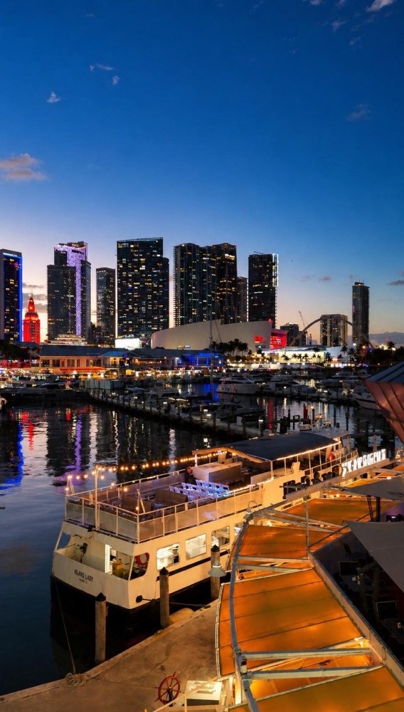 City skyline at dusk with illuminated buildings and a docked boat by the waterfront.