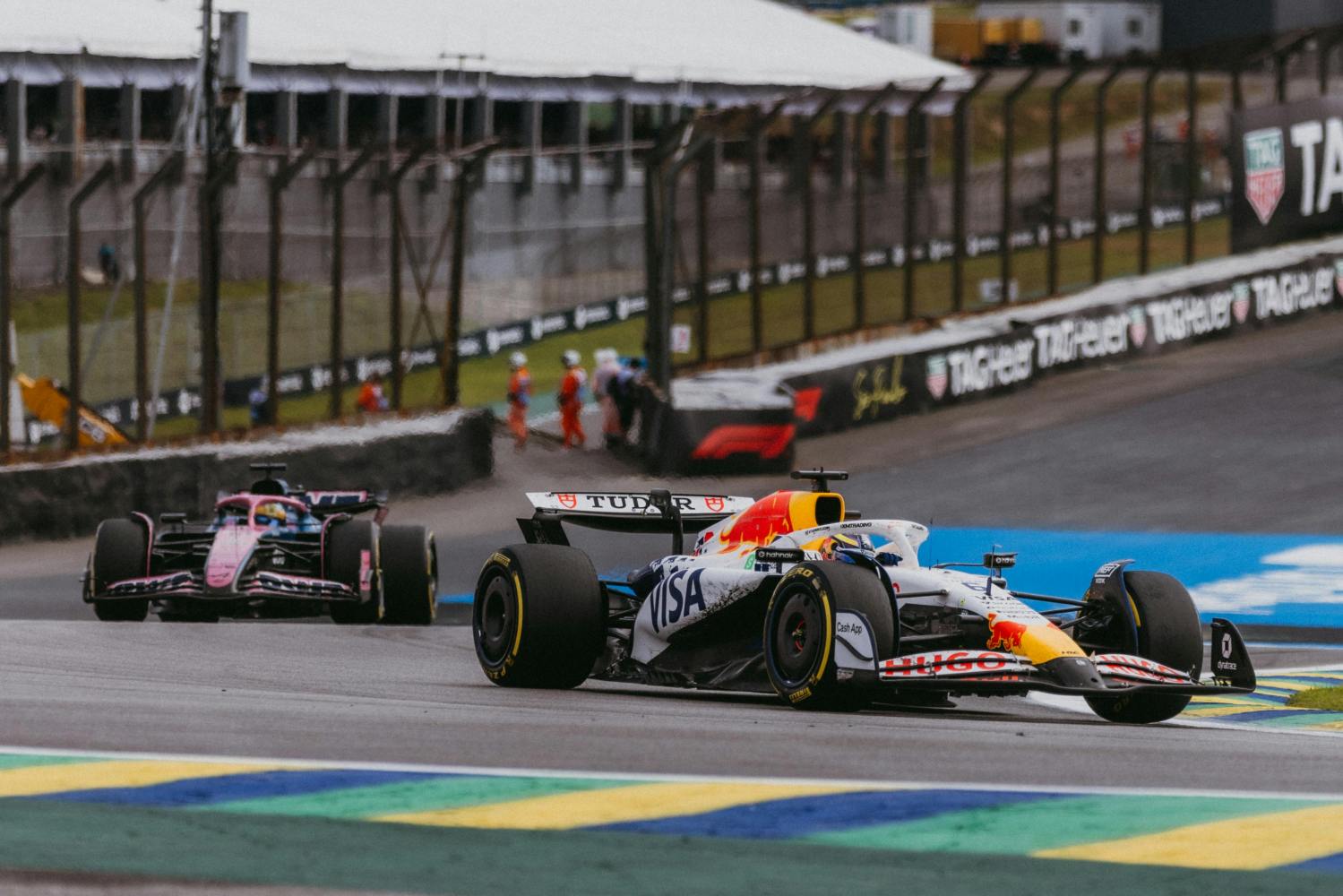 Two Formula 1 cars racing on a track with fencing and structures in the background.
