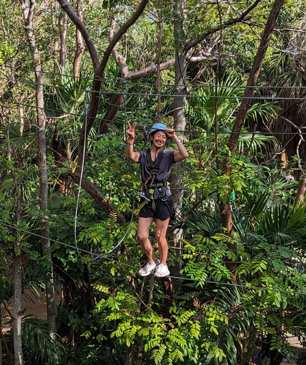 Person ziplining through lush green forest, wearing a helmet and harness.