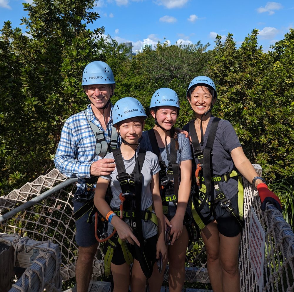 Four people in helmets and harnesses stand on a wooden platform surrounded by trees.