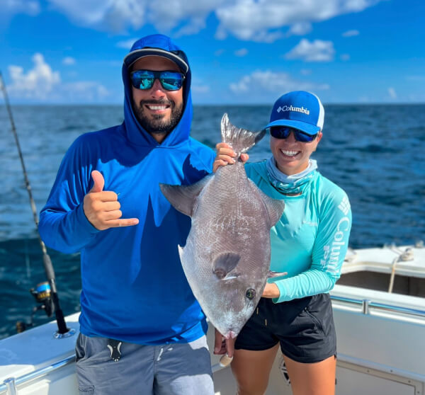 Two people on a boat holding a large fish, smiling under a blue sky.
